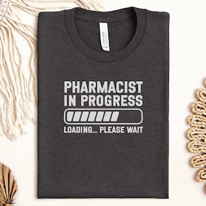 May include: Dark gray t-shirt with the text "PHARMACIST IN PROGRESS" in white, along with a loading bar graphic. Below the graphic, the text reads "LOADING... PLEASE WAIT". The shirt is folded neatly.