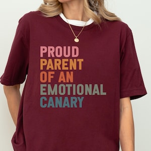 May include: A maroon t-shirt with the text "PROUD PARENT OF AN EMOTIONAL CANARY" in a stacked, colorful font. The shirt has short sleeves and a white crew neck collar.