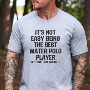 May include: A light gray t-shirt with the text "IT'S NOT EASY BEING THE BEST WATER POLO PLAYER BUT HERE I AM NAILING IT" in black. The shirt is short-sleeved.