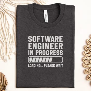 Könnte beinhalten: Dunkelgraues T-Shirt mit weißem Aufdruck "SOFTWARE ENGINEER IN PROGRESS", einem Ladebalken und den Worten "LOADING... PLEASE WAIT". Das Shirt hat einen Rundhalsausschnitt und ein "CANVAS"-Etikett.