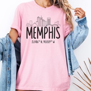 Puede incluir: Camiseta rosa claro con un diseño de horizonte negro y la palabra "MEMPHIS" en letras grandes. Debajo, el texto dice "35.1486° N, 90.0519° W". La camiseta está hecha de una tela suave y cómoda.