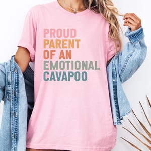May include: A pale pink t-shirt featuring the text "PROUD PARENT OF AN EMOTIONAL CAVAPOO" in a rainbow of colours. The shirt is worn with a blue denim jacket.