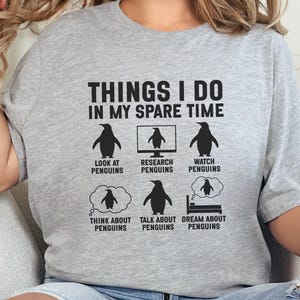 May include: A light gray t-shirt with the text "THINGS I DO IN MY SPARE TIME" and illustrations of penguins. The shirt shows penguins in various activities, including looking at, researching, and dreaming about penguins.
