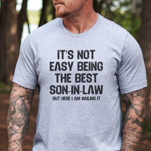 May include: A grey t-shirt with the text "IT'S NOT EASY BEING THE BEST SON-IN-LAW BUT HERE I AM NAILING IT" printed on the front.