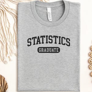 May include: A heather grey crew neck t-shirt featuring the black text "STATISTICS" in an arched collegiate font above the word "GRADUATE" enclosed in a black rectangular border. The shirt has a white neck label with the brand name Canvas.