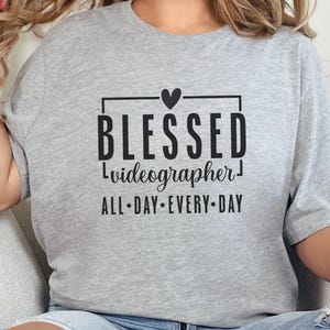 May include: A pale grey t-shirt featuring the text "BLESSED videographer ALL-DAY EVERY-DAY" in black, with a small heart above "BLESSED". The shirt has a relaxed fit.