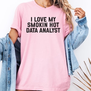 Peut inclure: T-shirt rose clair avec l'inscription noire "I LOVE MY SMOKIN HOT DATA ANALYST". La chemise est portée avec une veste en jean bleu clair. L'ensemble dégage un style décontracté.