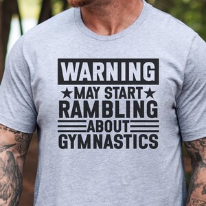 May include: A light grey t-shirt with the text "WARNING MAY START RAMBLING ABOUT GYMNASTICS" in a black, bold font. The text is arranged in a rectangular block. The shirt has short sleeves.