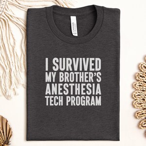 Könnte beinhalten: Dunkelgraues T-Shirt mit dem weißen Text: "I SURVIVED MY BROTHER'S ANESTHESIA TECH PROGRAM". Das T-Shirt ist gefaltet und liegt flach auf einer weißen Oberfläche. Es hat einen Rundhalsausschnitt und kurze Ärmel.