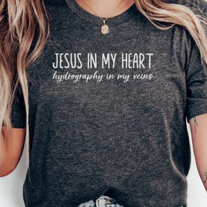 Peut inclure: T-shirt gris foncé avec l'inscription blanche "JESUS IN MY HEART" au-dessus de "hydrography in my veins". Le t-shirt a un col rond et des manches courtes. Un collier doré est également visible.