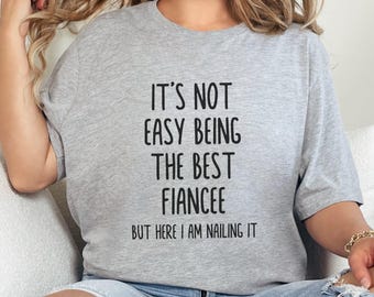 May include: Gray t-shirt with the text "It's not easy being the best fiancee but here I am nailing it."