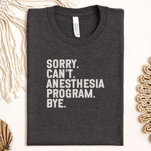 May include: Dark gray t-shirt with the text "SORRY. CAN'T. ANESTHESIA PROGRAM. BYE." in white block letters. The shirt is made of a soft material and has a crew neck. The shirt is folded neatly on a white surface.