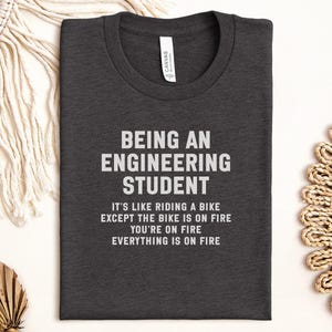 May include: Dark gray t-shirt with the text "BEING AN ENGINEERING STUDENT" in white. The shirt also has the quote: "IT'S LIKE RIDING A BIKE EXCEPT THE BIKE IS ON FIRE YOU'RE ON FIRE EVERYTHING IS ON FIRE."