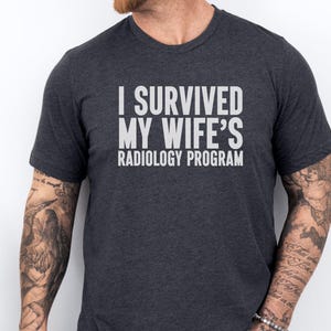 May include: A dark gray t-shirt with the text "I survived my wife's radiology program" printed in white.