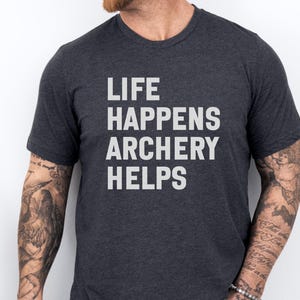 May include: Dark gray t-shirt with the text "LIFE HAPPENS ARCHERY HELPS" in white block letters. The shirt is a crew neck style and is being worn by a person with tattoos on their arms.