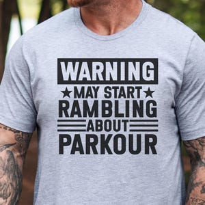 May include: A light gray t-shirt with the text "WARNING MAY START RAMBLING ABOUT PARKOUR" in a black, bold font. The text is arranged in a rectangular block. The shirt has short sleeves.