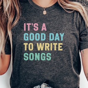 May include: A dark gray t-shirt with the text "It's a good day to write songs" in colorful letters. The text is in a rainbow color scheme, with pink, blue, yellow, and green.