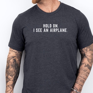 May include: A dark gray t-shirt with the text "Hold on. I see an airplane." printed in white.