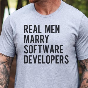 Peut inclure: Un t-shirt gris chiné à manches courtes et col rond avec le texte noir "REAL MEN MARRY SOFTWARE DEVELOPERS" imprimé en lettres capitales sans empattement au centre de la poitrine. Le vêtement est composé d'un tissu chiné doux.