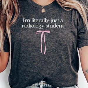 May include: Dark gray t-shirt with the text "I'm literally just a radiology student" in white. A pink bow graphic is below the text. The shirt is a crew neck style.