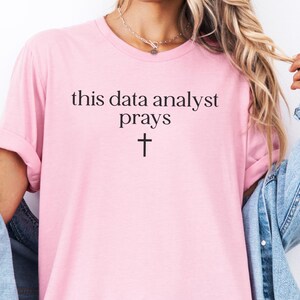 May include: A light pink short-sleeved t-shirt featuring the black text 'this data analyst prays' centered on the chest, positioned above a small black cross symbol. The shirt has a crew neckline and is worn with a blue denim jacket.