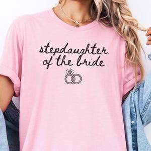 May include: A light pink t-shirt with the text "stepdaughter of the bride" in black script. Below the text are two interlocking rings with a diamond. The shirt is worn with a denim jacket.