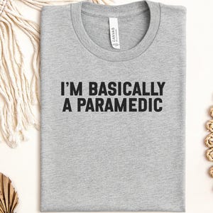 May include: A heather gray crew neck t-shirt featuring the black text "I'M BASICALLY A PARAMEDIC" printed on the chest. The shirt has a standard short-sleeve design with a visible brand label inside the collar area, showcasing a simple, clean aesthetic.