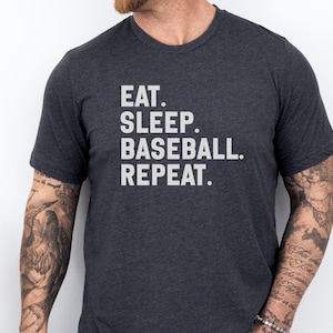 May include: Dark gray t-shirt with the words "EAT. SLEEP. BASEBALL. REPEAT." printed in white. The shirt is a crew neck style and appears to be made of a soft material. The image shows the shirt on a person.
