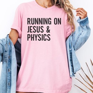 May include: A light pink t-shirt with the text "RUNNING ON JESUS & PHYSICS" in bold black letters. The shirt has short sleeves and a crew neck. A light blue denim jacket is draped over the shoulders.