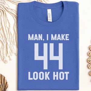 May include: A blue t-shirt with the text "MAN, I MAKE 44 LOOK HOT" in white. The shirt is folded and lies on a white surface, with decorative elements in the background.