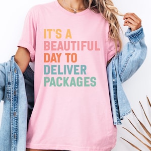 May include: Pink t-shirt with the text "IT'S A BEAUTIFUL DAY TO DELIVER PACKAGES" in orange, pink, orange, teal, and green. The shirt is worn with a denim jacket.