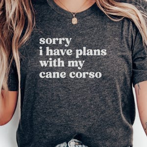 Puede incluir: Camiseta gris oscuro con el texto blanco "sorry i have plans with my cane corso". La camiseta es de cuello redondo y tiene mangas cortas. La imagen muestra la parte superior del cuerpo de una persona que lleva la camiseta.