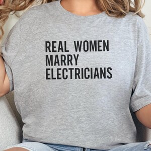 Op de afbeelding: Lichtgrijs t-shirt met de tekst "REAL WOMEN MARRY ELECTRICIANS" in vette zwarte letters. Het shirt heeft korte mouwen en een relaxte pasvorm. De tekst staat centraal op de voorkant van het kledingstuk.