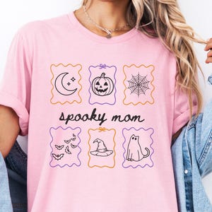 May include: A light pink t-shirt featuring a graphic print with the text "spooky mom" in black script. The design includes six rectangular frames containing black line art of a crescent moon, a jack-o'-lantern, a spider web, bats, a witch hat, and a ghost.