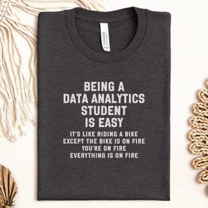 May include: Charcoal grey t-shirt with white text: "BEING A DATA ANALYTICS STUDENT IS EASY". The shirt also has the phrases "IT'S LIKE RIDING A BIKE EXCEPT THE BIKE IS ON FIRE YOU'RE ON FIRE EVERYTHING IS ON FIRE."