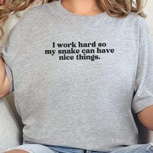 May include: A pale grey t-shirt featuring the black text: "I work hard so my snake can have nice things". The shirt has a crew neck and short sleeves.