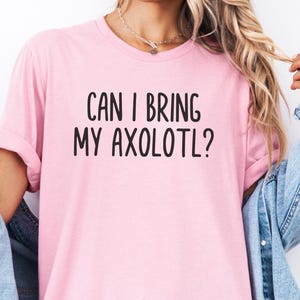 Könnte beinhalten: Hellrosa T-Shirt mit dem schwarzen, fettgedruckten Text "CAN I BRING MY AXOLOTL?". Das T-Shirt hat kurze Ärmel und einen Rundhalsausschnitt. Eine Jeansjacke wird darüber getragen.