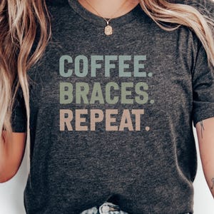 Puede incluir: Camiseta gris oscuro con el texto "COFFEE. BRACES. REPEAT." en una fuente multicolor. La camiseta es de cuello redondo y tiene mangas cortas. La imagen muestra la parte superior del cuerpo de una persona que lleva la camiseta.