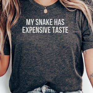 May include: A dark charcoal grey short-sleeved t-shirt featuring the white text "MY SNAKE HAS EXPENSIVE TASTE" printed in a clean, sans-serif font across the chest. The garment is made of a heathered fabric with a visible texture.