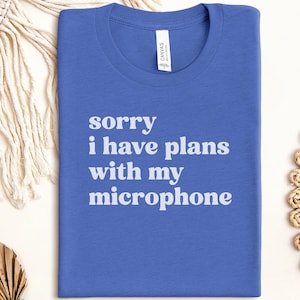 Puede incluir: Una camiseta azul con el texto "sorry I have plans with my microphone" en blanco. La camiseta está hecha de un material suave y probablemente sea de corte unisex. La camiseta está doblada cuidadosamente sobre una superficie blanca.