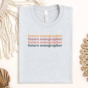 Puede incluir: Una camiseta gris con el texto "future sonographer" repetido cuatro veces en diferentes colores. El texto está en un estilo de fuente retro.