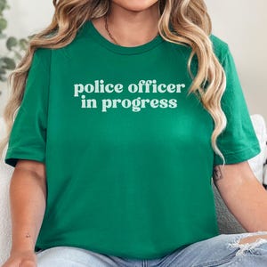 May include: A green t-shirt with the text "police officer in progress" in white, centered on the chest. The shirt has short sleeves and a crew neck. The person is wearing blue jeans.