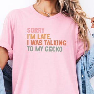 Könnte beinhalten: Hellrosa T-Shirt mit dem Text "SORRY I'M LATE, I WAS TALKING TO MY GECKO" in einem Retro-Schriftzug. Das Shirt hat hochgekrempelte Ärmel und wird mit einer Jeansjacke getragen.