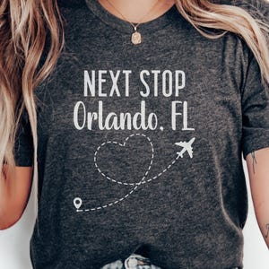 Puede incluir: Camiseta gris oscuro con el texto "NEXT STOP Orlando, FL" en blanco. Una línea de puntos forma un corazón y un avión. Una camiseta con temática de viajes.