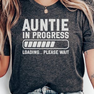 May include: A dark charcoal grey short-sleeve t-shirt featuring white text that reads AUNTIE IN PROGRESS above a loading bar graphic and the text LOADING... PLEASE WAIT. The shirt has a crew neckline and a standard fit, made of a soft fabric.