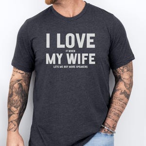 May include: Dark gray t-shirt with the text "I LOVE IT WHEN MY WIFE LETS ME BUY MORE SPEAKERS" in white block letters. The shirt is a crew neck style and is being worn by a person.