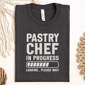May include: Dark gray t-shirt with the words "PASTRY CHEF IN PROGRESS" in white block letters. Below the text is a loading bar graphic and the words "LOADING... PLEASE WAIT". The shirt is folded and displayed on a white surface.