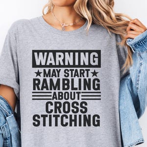 May include: A light grey t-shirt featuring the black text: "WARNING MAY START RAMBLING ABOUT CROSS STITCHING". The text is in a rectangular block with a star and lines. The shirt has short sleeves and is casual wear.