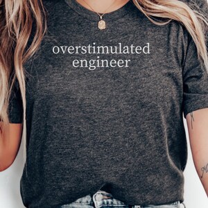 May include: Dark grey crew neck t-shirt with the text "overstimulated engineer" in white. The shirt is made of a soft material. The image shows the upper body of a person wearing the t-shirt. The person has a tattoo on their arm.