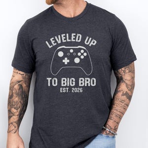 Könnte beinhalten: Dunkelgraues T-Shirt mit dem Text "LEVELED UP TO BIG BRO EST. 2026" und einer weißen Controller-Grafik. Das Shirt besteht aus einem weichen Material und eignet sich für Freizeitkleidung. Ein tolles Geschenk für einen neuen großen Bruder.